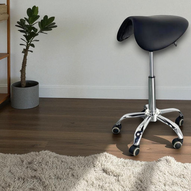 Black saddle-style stool with wheels on a wooden floor next to a plant and rug.